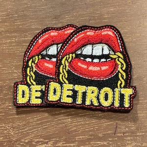 May include: Embroidered patch featuring two sets of red lips with gold chain detailing. The patch has a black border and the words "DE' DETROIT" in yellow lettering.