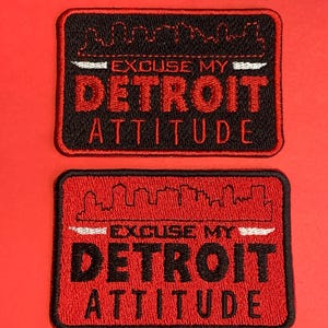 May include: Two rectangular embroidered patches with the text "EXCUSE MY DETROIT ATTITUDE". The patches are black with red borders and feature a skyline design above the text. The background is red.