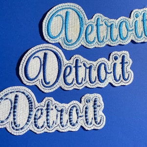May include: Three embroidered patches featuring the word Detroit in a cursive script font. The top patch has light blue thread, the middle patch has medium blue thread, and the bottom patch has dark blue thread, all on a white background.
