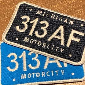 May include: Two embroidered patches in the shape of license plates. One is navy blue with white text that reads "MICHIGAN 313AF MOTORCITY". The other is light blue with the same text.