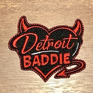 May include: Embroidered patch in a heart shape with a devilish design. The patch features the words "Detroit Baddie" in red script over a black background, outlined in red. A red devil tail and horns complete the design.
