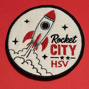 May include: A circular embroidered patch with a white background and a black border. It features a white rocket with red accents, black smoke, and stars. The text reads Rocket CITY HSV in black and red lettering with small black stars.