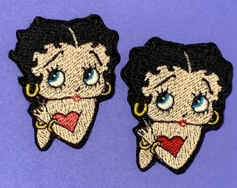 May include: Two embroidered patches featuring the cartoon character Betty Boop. Each patch has black hair, blue eyes, a red heart-shaped mouth, and gold hoop earrings. The patches are on a purple background.