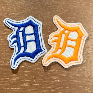 May include: Two embroidered patches in the shape of the letter D, featuring a gothic-style font. One patch has a blue border and blue stitching, while the other has an orange border and orange stitching. Both patches have a white fabric center.