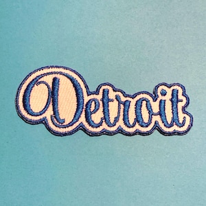 May include: Embroidered patch featuring the word "Detroit" in a cursive font. The text is in blue, outlined with a white border, and a dark blue trim. The patch is on a light blue background.