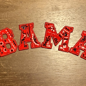 May include: Red embroidered patches spelling "BAMA". Each letter is outlined in red and filled with a paisley pattern in red, black, and white. The patches are arranged on a wooden surface.