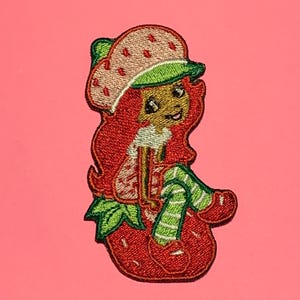 May include: Embroidered patch featuring a cartoon character with red hair, a strawberry hat, and green and white striped pants. The character is sitting on a large red strawberry. The patch has a pink background.