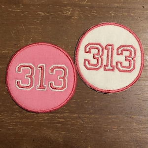 May include: Two circular embroidered patches featuring the number 313 in a bold, block-style font. One patch has a pink background with a red border, while the other has an off-white background with a red border. Both patches have red stitching.