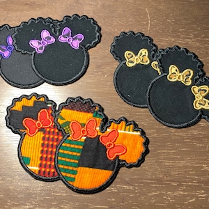 May include: Black fabric patches shaped like a mouse head with a bow. The bows are purple, gold, and red. The mouse head has a black afro. The patches have a black border and are on a brown surface.