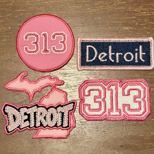 May include: Four embroidered patches featuring Detroit themes. One circular pink patch with white 313 text, one rectangular navy patch with white Detroit text, one pink Michigan state outline patch, and one pink 313 patch with white text.