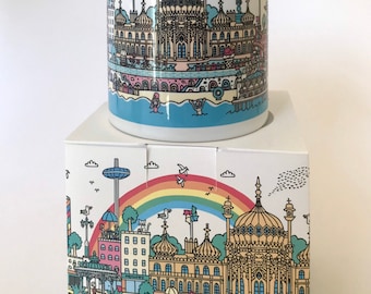 May include: A white ceramic mug and its matching square cardboard box, both featuring a colorful, detailed illustration of the Brighton Pavilion, a rainbow, beach huts, and people swimming in the sea under a clear sky with birds.