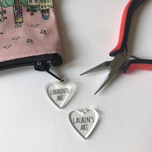 Pu&ograve; includere: Due ciondoli a forma di cuore in acrilico trasparente con il testo "Lauren's Art" su ciascuno. I ciondoli sono su una superficie bianca con un paio di pinze rosse e nere.