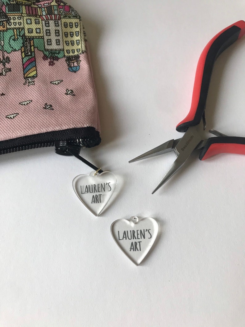 Pu&ograve; includere: Due ciondoli a forma di cuore in acrilico trasparente con il testo "Lauren's Art" su ciascuno. I ciondoli sono su una superficie bianca con un paio di pinze rosse e nere.