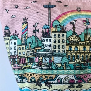 Pu&ograve; includere: Una pochette con cerniera in tela rosa con un'illustrazione colorata di una citt&agrave; sul mare. L'illustrazione include edifici, una ruota panoramica, un arcobaleno e una scena di spiaggia.