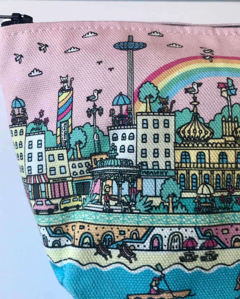 Pu&ograve; includere: Una pochette con cerniera in tela rosa con un'illustrazione colorata di una citt&agrave; sul mare. L'illustrazione include edifici, una ruota panoramica, un arcobaleno e una scena di spiaggia.