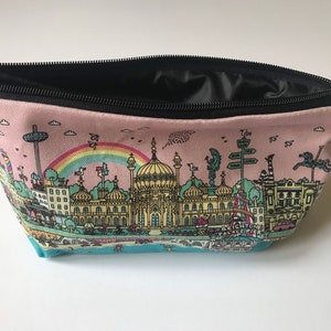 Pu&ograve; includere: Una pochette con zip in tela rosa con una cerniera nera e un'illustrazione colorata del Brighton Palace Pier sul davanti. L'illustrazione include un arcobaleno, edifici e il testo "Brighton Palace Pier".