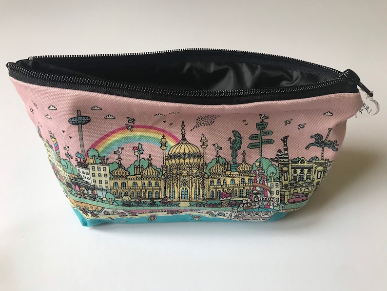 Pu&ograve; includere: Una pochette con zip in tela rosa con una cerniera nera e un'illustrazione colorata del Brighton Palace Pier sul davanti. L'illustrazione include un arcobaleno, edifici e il testo "Brighton Palace Pier".