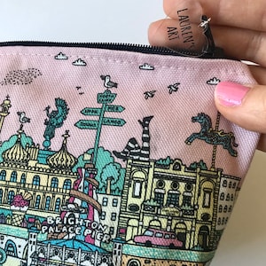 Pu&ograve; includere: Una pochette rosa in tela con cerniera, con un'illustrazione colorata del Brighton Palace Pier. La pochette ha un'etichetta di plastica trasparente a forma di cuore con il testo "Lauren's Art" su di essa.