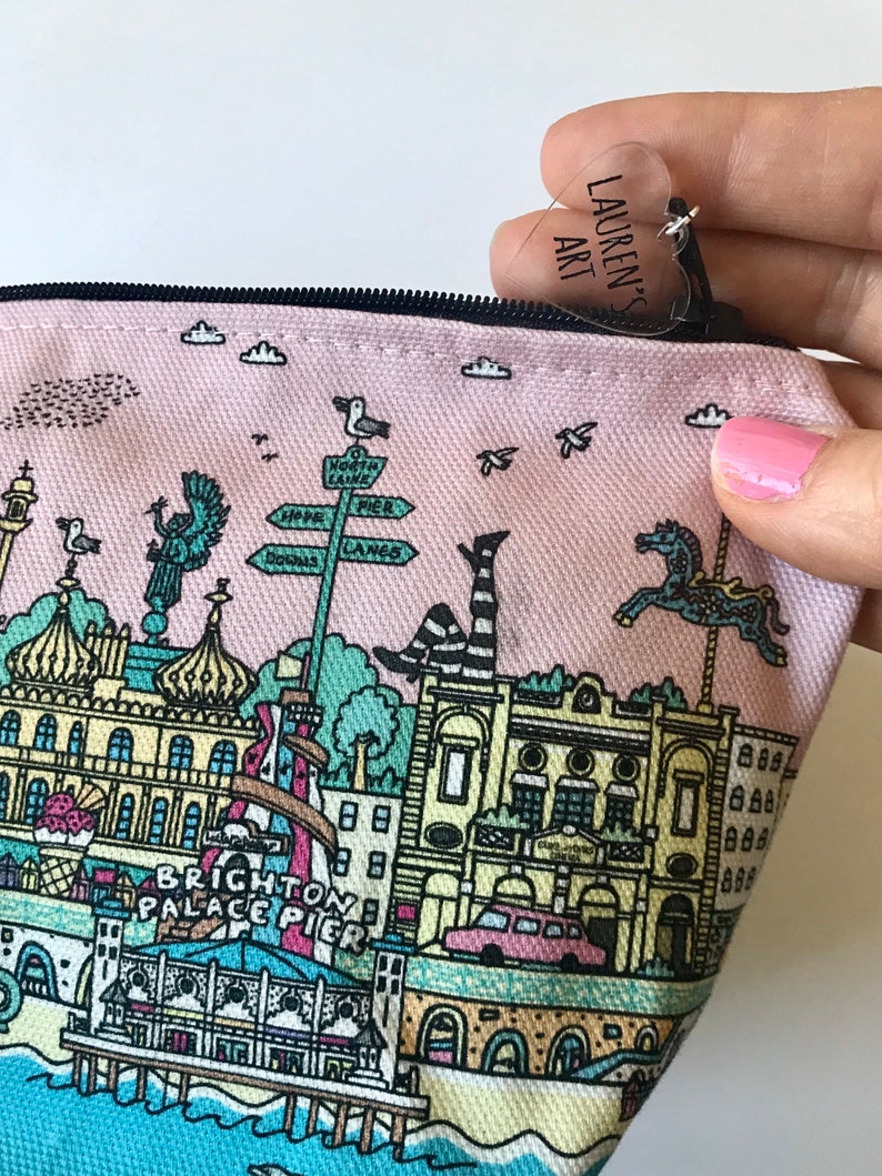 Pu&ograve; includere: Una pochette rosa in tela con cerniera, con un'illustrazione colorata del Brighton Palace Pier. La pochette ha un'etichetta di plastica trasparente a forma di cuore con il testo "Lauren's Art" su di essa.