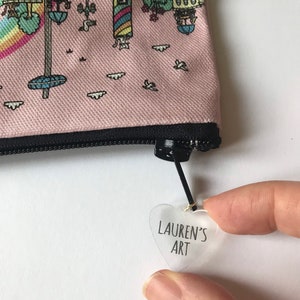 Pu&ograve; includere: Una pochette rosa con cerniera con una scena di citt&agrave; fantasiosa stampata su di essa. La pochette ha un'etichetta trasparente a forma di cuore attaccata al tiretto della cerniera con il testo "LAUREN'S ART" stampato su di essa.