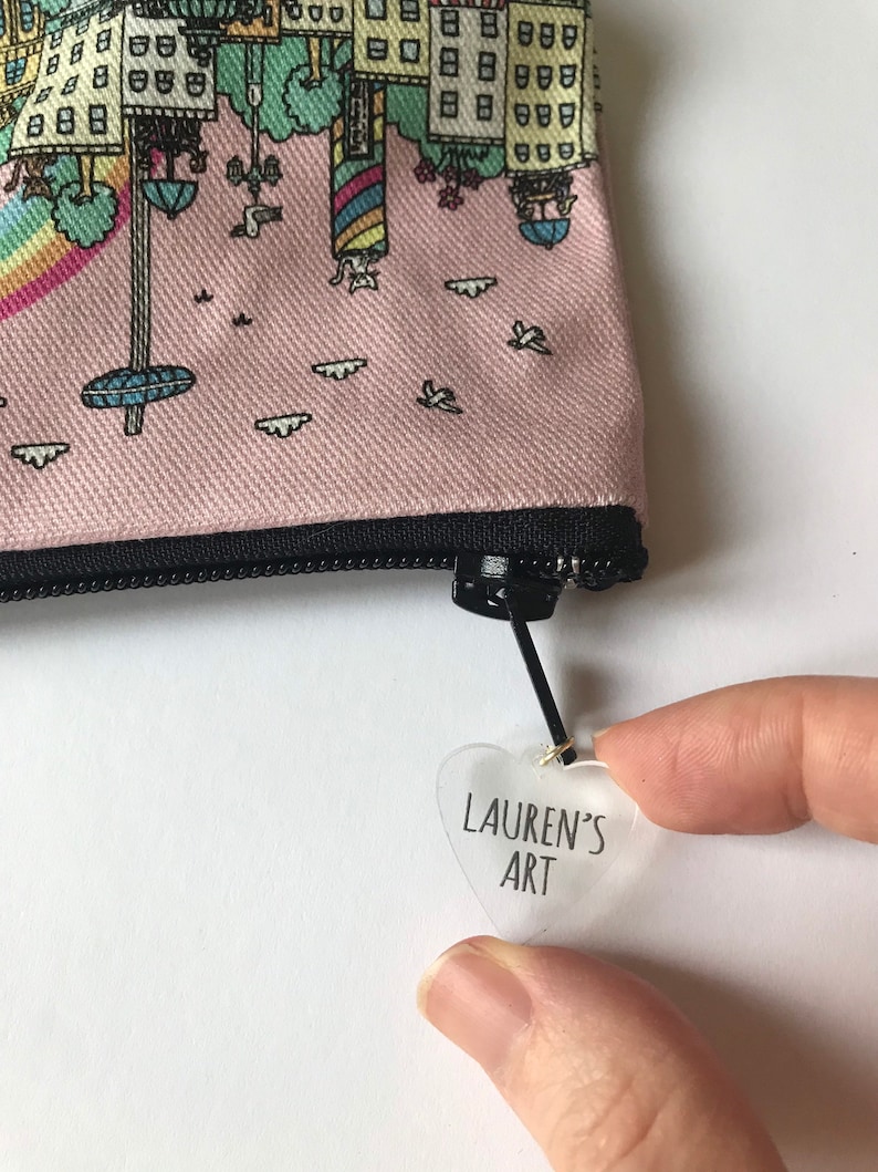 Pu&ograve; includere: Una pochette rosa con cerniera con una scena di citt&agrave; fantasiosa stampata su di essa. La pochette ha un'etichetta trasparente a forma di cuore attaccata al tiretto della cerniera con il testo "LAUREN'S ART" stampato su di essa.