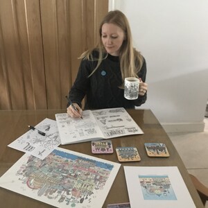 Pu&ograve; includere: Una donna &egrave; seduta a un tavolo e sta lavorando a un disegno dettagliato di una citt&agrave;. Sta tenendo una tazza con un disegno di una citt&agrave;. Ci sono diverse stampe della citt&agrave; sul tavolo, insieme a sottobicchieri.