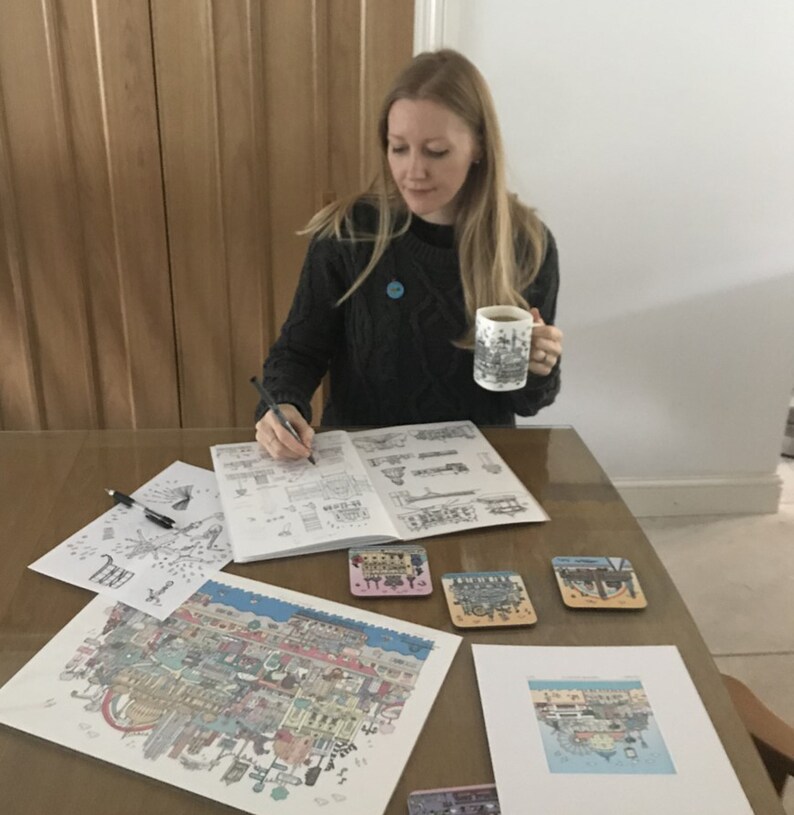 Pu&ograve; includere: Una donna &egrave; seduta a un tavolo e sta lavorando a un disegno dettagliato di una citt&agrave;. Sta tenendo una tazza con un disegno di una citt&agrave;. Ci sono diverse stampe della citt&agrave; sul tavolo, insieme a sottobicchieri.