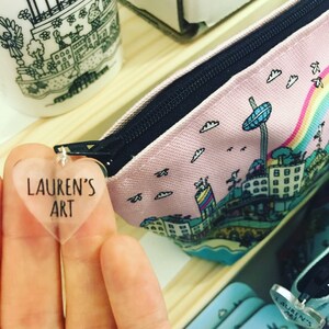 Pu&ograve; includere: Una pochette rosa con cerniera con un design di citt&agrave; con skyline colorato. La pochette &egrave; aperta e un portachiavi trasparente a forma di cuore con il testo "Lauren's Art" &egrave; attaccato alla cerniera.