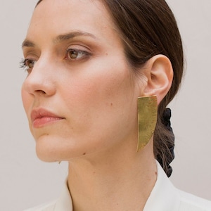 May include: A woman wearing a white collared shirt with a black harness and a gold circular pendant. She is also wearing large, rectangular gold earrings. The background is a plain, light-coloured wall.