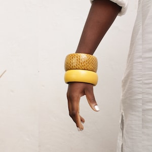 Può includere: Un set di due braccialetti, uno giallo e uno marrone. I braccialetti sono impilati sul polso di una persona.