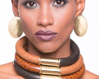 Op de afbeelding: Een vrouw draagt een gelaagde lederen choker met gouden accenten. De ketting is bruin en zwart en heeft een tribale uitstraling. De vrouw draagt ook grote gouden oorringen.