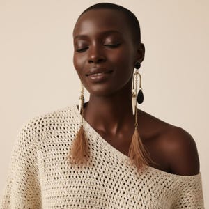 May include: Long, dangling earrings with a gold chain, black and white accents, and brown feathers. The earrings are worn by a person wearing a cream-coloured, open-weave sweater.