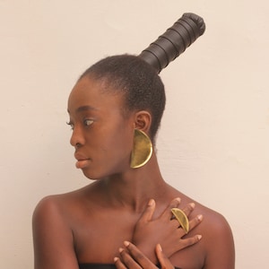 May include: A woman with dark brown skin is wearing large gold half-circle earrings and a gold ring. Her hair is styled in a high bun with a black leather hair tie.
