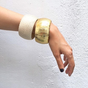 Könnte beinhalten: Zwei Armbänder, die auf einem Handgelenk übereinander liegen. Das untere Armband ist golden mit einem strukturierten Design. Das obere Armband ist weiß mit einem Schlangenhautmuster.