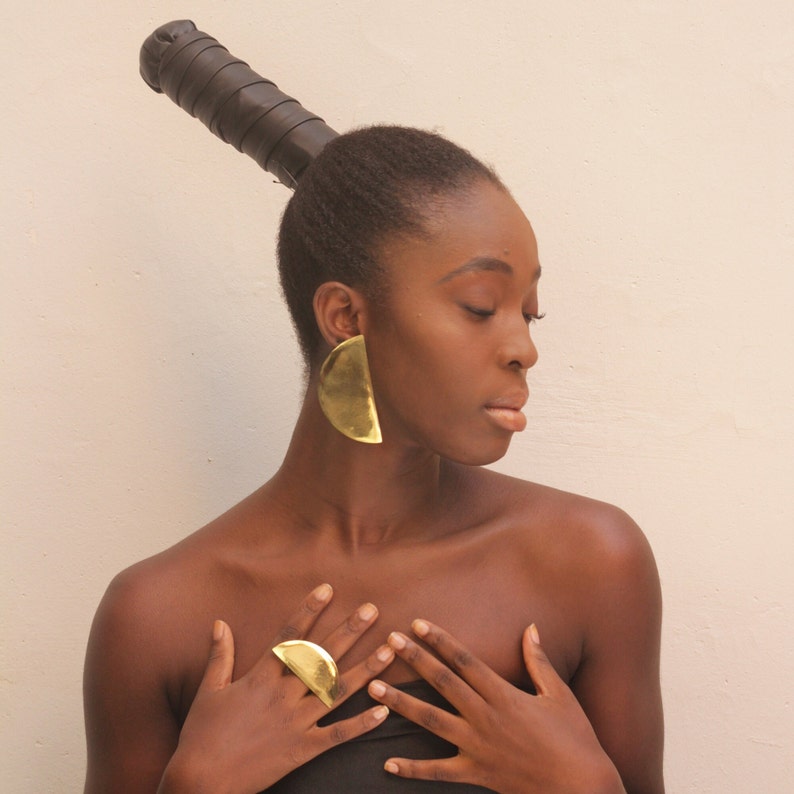 May include: A woman with dark brown hair styled in a high ponytail with a black leather hair tie is wearing large gold half-circle earrings and a gold ring on her finger.