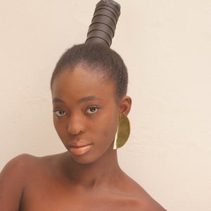 May include: A woman with dark brown hair styled in a high bun with a black hair tie. She is wearing large gold hoop earrings.