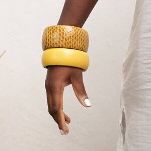 Könnte beinhalten: Zwei Holzarmreifen an einer dunkelhäutigen Hand. Der obere Armreif hat eine strukturierte, hellbraune Oberfläche, während der untere ein glattes, gelbes Vollton ist. Die Armreifen sind gestapelt und zeigen einen Kontrast in Textur und Farbe. Modeaccessoire.