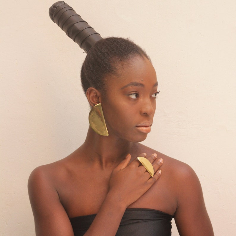 May include: A woman with dark brown hair styled in a high bun with a black leather band. She is wearing large gold half-circle earrings and a gold ring on her right hand.