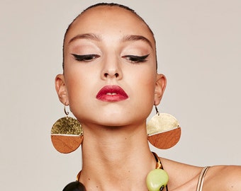 Op de afbeelding: Een vrouw met kort, geschoren haar draagt grote, ronde oorbellen met een gouden en bruin lederen ontwerp. Ze draagt ook een veelkleurige kralenketting.