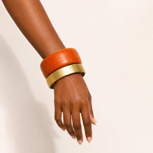 May include: Two stacked bangle bracelets worn on the wrist. The top bangle is a wide, textured orange band with a grid-like pattern. The bottom bangle is a thinner, smooth metallic gold band with a subtle horizontal lined texture across the surface.