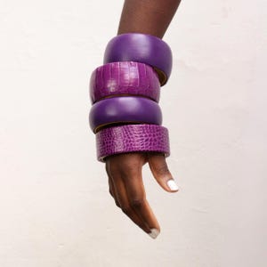 Può includere: Quattro braccialetti viola impilati su un braccio. Due sono lisci e due hanno un motivo strutturato a forma di pelle di coccodrillo. I braccialetti sono in diverse tonalità di viola, creando un accessorio audace.