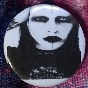 May include: Black and white button featuring a portrait of a person with dark hair and dark eye makeup. The person is wearing a black shirt.