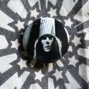 May include: Black and white button featuring a portrait of a person wearing a tall, cylindrical hat with a striped pattern. The person's face is partially obscured by the hat and has a serious expression.