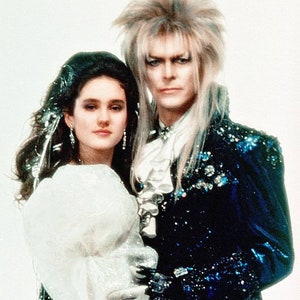 May include: A man and a woman in a formal setting. The man has long, silver hair and is wearing a blue velvet jacket with sequins. The woman is wearing a white dress with a large, white bow.