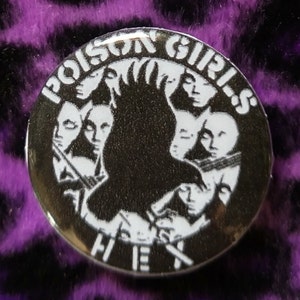 May include: A silver button badge with a black and white image of a crow with a circle of faces around it. The text "Poison Girls" is written in a circle around the crow and the text "Hex" is written below the crow.
