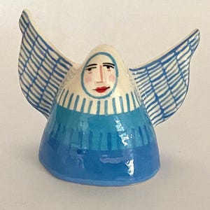 May include: A ceramic figurine of an angel with a bell-shaped body in shades of blue. The figure features a painted face with red lips, a white head covering, and large, outstretched wings decorated with a blue and white grid pattern.