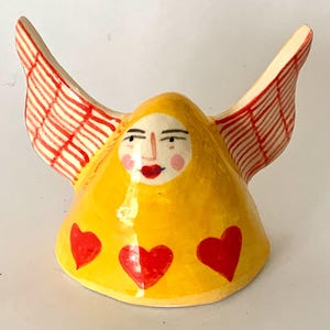 May include: A quirky ceramic ornament depicting a figure with wings. The figure is predominantly yellow, featuring a face and three red hearts. The wings are white with red stripes. The face has rosy cheeks and red lips.