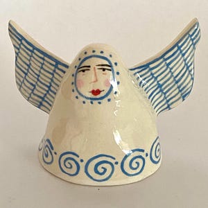 May include: A cream-coloured ceramic figurine shaped like a bell with outstretched wings. The piece features a hand-painted face with red lips and pink cheeks, surrounded by blue dots. Blue grid patterns adorn the wings, and blue swirls decorate the base.
