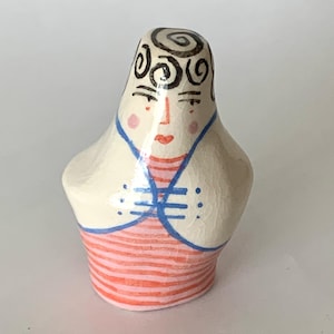 May include: A ceramic figurine of a woman with a cream-coloured body, red and orange striped bottom, and a blue and white top. The figurine has black swirled hair, pink cheeks, and red lips.