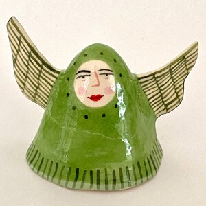 May include: A ceramic figurine shaped like a bell with a green hood and two outstretched wings. The face features painted eyes, a nose, pink cheeks, and red lips. The hood has black dots, and the base and wings have green striped patterns.
